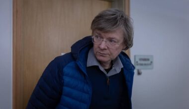 En person står i en døråpning i et rom med lyse vegger og en tre-dør. Ved siden av døren er det et skilt. Personen har på seg en blå jakke og en lys skjorte. Omgivelsene fremstår som nøytrale, uten synlige detaljer som kan gi mer kontekst.