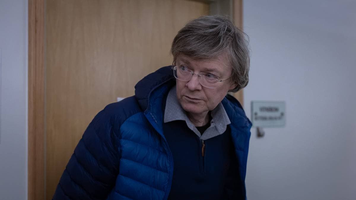 En person står i en døråpning i et rom med lyse vegger og en tre-dør. Ved siden av døren er det et skilt. Personen har på seg en blå jakke og en lys skjorte. Omgivelsene fremstår som nøytrale, uten synlige detaljer som kan gi mer kontekst.