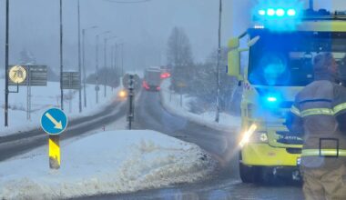 Redningsmannskap står ved en avstengt vei, omringet av snødekte omgivelser. En brannbil med blålys er parkert på veien, mens en lastebil sirkulerer i bakgrunnen. Det er dårlig sikt på grunn av snøvær, og veien er dekket av snø. Veiskilt og lamper er synlige langs veien.