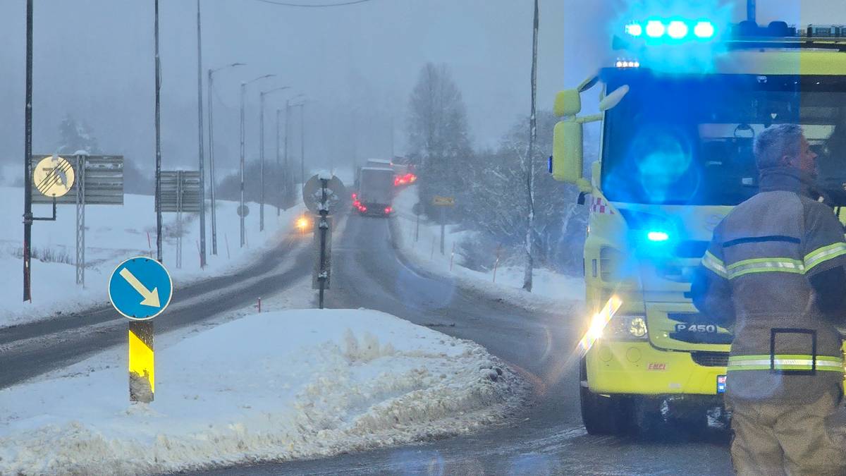 Redningsmannskap står ved en avstengt vei, omringet av snødekte omgivelser. En brannbil med blålys er parkert på veien, mens en lastebil sirkulerer i bakgrunnen. Det er dårlig sikt på grunn av snøvær, og veien er dekket av snø. Veiskilt og lamper er synlige langs veien.