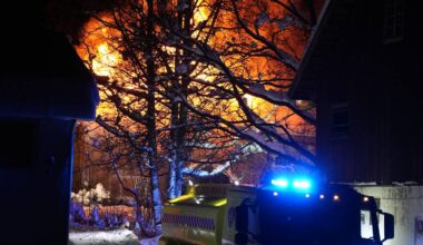 En stor brann lyser opp natten bak trærne, med flammer som stiger fra en bygning. Et brannbil står parkert i snøen, opplyst av blålys. Brannmannskap arbeider i nærheten, mens røyken stiger fra brannstedet. Omgivelsene er dekket av snø, og flere bygninger er delvis synlige.
