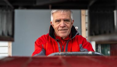 Bildet viser en person med grått hår iført en rød jakke og svart hette, som sitter bak en ratt. Omgivelsene inkluderer en bakgrunn med en grå vegg og en dør. Personen ser ut til å være i ferd med å kjøre eller styre et kjøretøy, muligens en gaffeltruck. En rød overflate er synlig foran personen, noe som kan indikere en arbeidsplass. (Bildebeskrivelsen er laget av en KI-tjeneste)