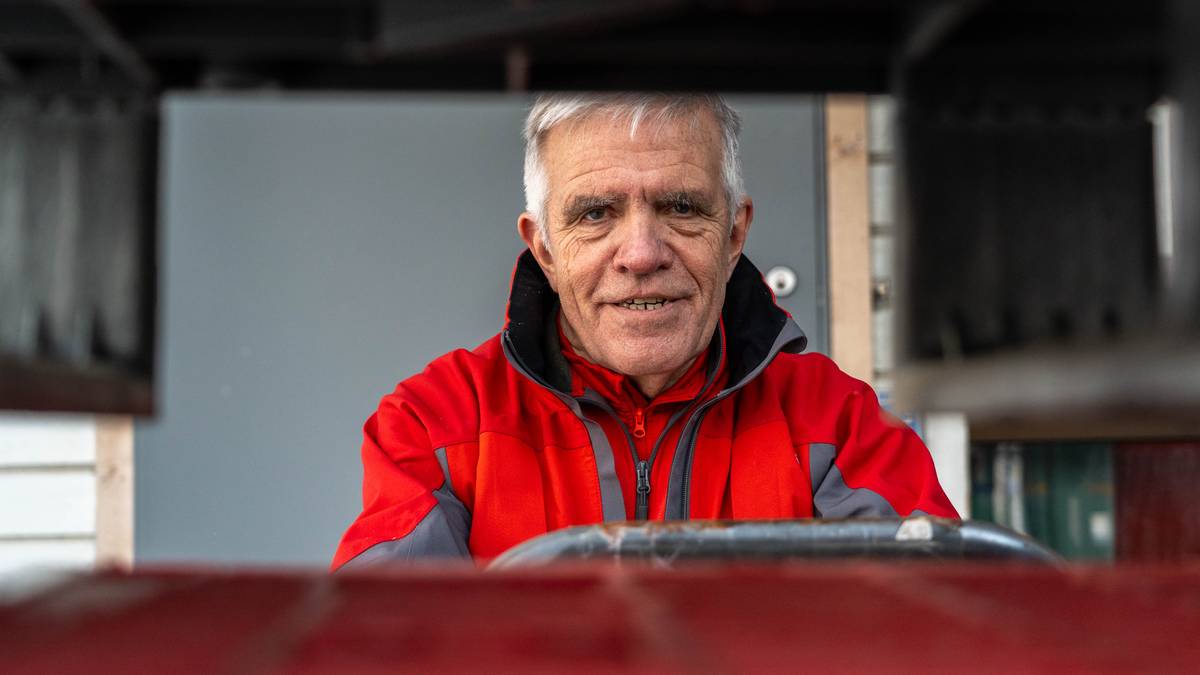 Bildet viser en person med grått hår iført en rød jakke og svart hette, som sitter bak en ratt. Omgivelsene inkluderer en bakgrunn med en grå vegg og en dør. Personen ser ut til å være i ferd med å kjøre eller styre et kjøretøy, muligens en gaffeltruck. En rød overflate er synlig foran personen, noe som kan indikere en arbeidsplass. (Bildebeskrivelsen er laget av en KI-tjeneste)