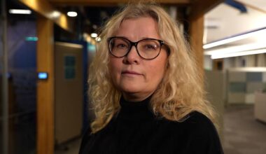 En person med krøllete hår står i en moderne kontoromgivelser. Bakgrunnen består av glatte, lyse vegger og møbler. Belysningen er dempet, men det er flere lyskilder synlige i taket. Personen har på seg en svart genser og ser direkte mot kameraet. (Bildebeskrivelsen er laget av en KI-tjeneste)