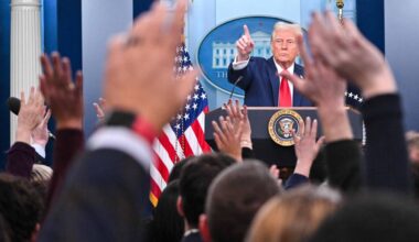 En person står bak en talerstol i et offisielt rom, med det amerikanske flagget i bakgrunnen. Hender fra en gruppe mennesker rekker opp i luften, noe som indikerer spørsmål eller respons. Rommet har et formelt preg med søyler og blå vegger. En offisiell presidentsegl er synlig på talerstolen. (Bildebeskrivelsen er laget av en KI-tjeneste)