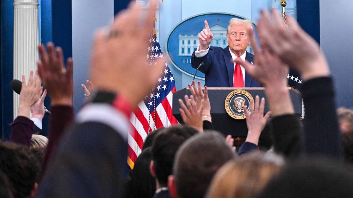 En person står bak en talerstol i et offisielt rom, med det amerikanske flagget i bakgrunnen. Hender fra en gruppe mennesker rekker opp i luften, noe som indikerer spørsmål eller respons. Rommet har et formelt preg med søyler og blå vegger. En offisiell presidentsegl er synlig på talerstolen. (Bildebeskrivelsen er laget av en KI-tjeneste)