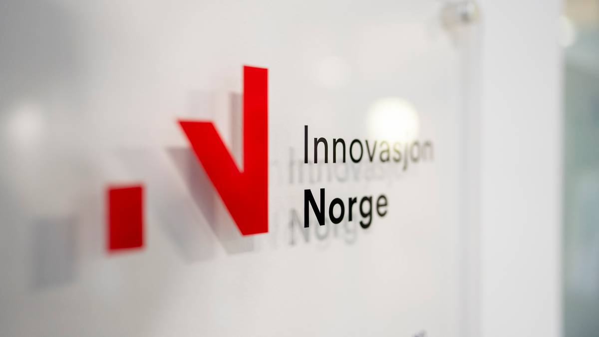 Bildet viser et nærbilde av en vegg med et skilt for "Innovasjon Norge". Skiltet har en rød "V" og teksten "Innovasjon Norge" i svart. Det er en lys bakgrunn, og detaljer på veggen er uskarpe. Omgivelsene gir inntrykk av et moderne kontorlandskap.