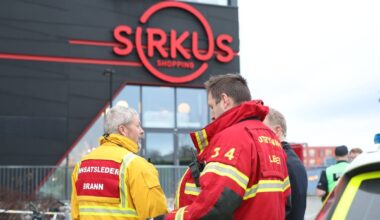 Brannmannskap står utenfor Sirkus Shopping, hvor de diskuterer situasjonen. To brannmenn er kledd i uniformer med refleksstriper og identifikasjon. Omgivelsene viser en moderne bygning med store vinduer. Politifolk kan sees i bakgrunnen, noe som indikerer en koordinert respons.