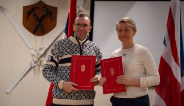 To personer står foran en skjerm med nasjonale flagg i bakgrunnen. Begge holder røde dokumenter med gullmotiv. Den ene er kledd i en mønstrete genser, mens den andre har på seg en lysere genser. Veggen bak dem har dekorerte sverd og et skjold.