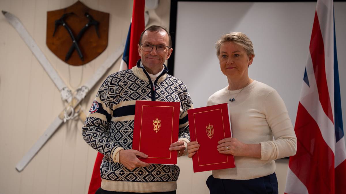 To personer står foran en skjerm med nasjonale flagg i bakgrunnen. Begge holder røde dokumenter med gullmotiv. Den ene er kledd i en mønstrete genser, mens den andre har på seg en lysere genser. Veggen bak dem har dekorerte sverd og et skjold.