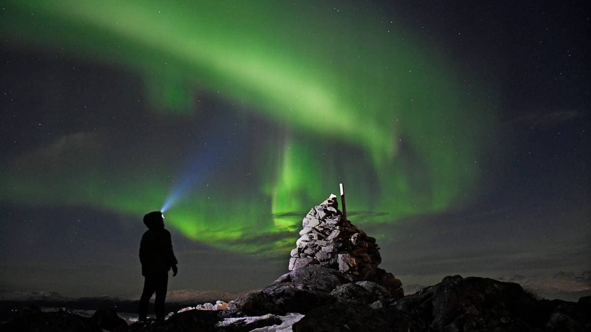 En person står på en fjellknaus med hodet hevet mot nordlyset som lyser opp nattehimmelen. Personen har på seg en jakke og har en lommelykt som lyser mot lyset. I bakgrunnen er det en steinpile, og stjernene er synlige på den mørke himmelen. Nordlyset skaper en dramatisk grønnfarge som dominerer scenen. (Bildebeskrivelsen er laget av en KI-tjeneste)