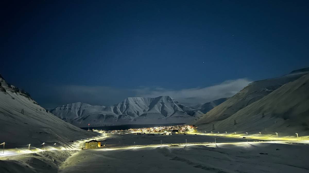 En naturskjønn utsikt viser en snødekt dal under stjerneklar himmel. I dalbunnen lyser en liten bosetning opp med gatelys, omringet av majestetiske fjell. Snøen dekker terrenget, og en vei er synlig som slynger seg ned mot bygningene. Området gir inntrykk av avsides beliggenhet og vinterlige forhold. (Bildebeskrivelsen er laget av en KI-tjeneste)