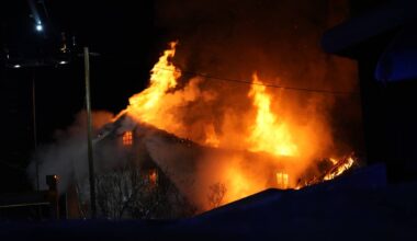 En bygning er sterkt i brann med store flammer som stiger opp fra taket. Røyk fyller luften rundt bygningen, og lysene fra brannkjøretøy kan sees i bakgrunnen. Det er mørkt, noe som forsterker synligheten av ilden. Omgivelsene er snødekte, noe som indikerer vinterforhold.