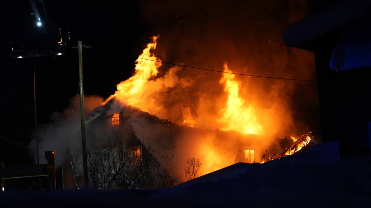 En bygning er sterkt i brann med store flammer som stiger opp fra taket. Røyk fyller luften rundt bygningen, og lysene fra brannkjøretøy kan sees i bakgrunnen. Det er mørkt, noe som forsterker synligheten av ilden. Omgivelsene er snødekte, noe som indikerer vinterforhold.