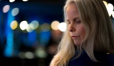 Bildet viser en person med langt, lyst hår, som står delvis vendt mot kameraet. Bakgrunnen er uklar, med flere svake lysglimt og en blålig fargetone, noe som kan indikere en sosial setting. Det er få detaljer synlige, men omgivelsene gir inntrykk av en sammenkomst eller et arrangement. Det er ingen grafiske elementer eller spesifikke objekter i bildet som skiller seg ut. (Bildebeskrivelsen er laget av en KI-tjeneste)