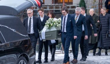 En gruppe personer bærer en kiste, dekorert med blomster, ut fra en kirke. De fleste av dem er ikledd formelle klær, og handlingen skjer på en vinterdag med snø på bakken. I bakgrunnen kan man se en svart bil, antagelig en begravelsesbil, parkert ved inngangen. En del tilskuere står på siden av veien og observerer bevegelsene. (Bildebeskrivelsen er laget av en KI-tjeneste)