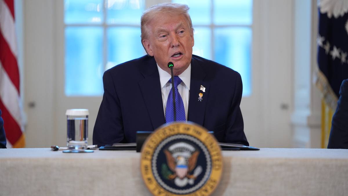 Bilde av en person som sitter ved et bord, iført mørk dress og blå slips, med en vannflaske foran seg. Det er et offisielt rom med vinduer i bakgrunnen og flagg på veggen. Bordet har en presidentemblem plassert sentralt. En annen person er delvis synlig til høyre.