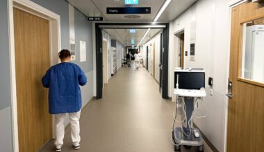 En ansatt i helsesektoren i blå arbeidsdrakt står med ryggen til foran en lukket dør i en sykehuskorridor. I bakgrunnen er flere personer i hvite klær som beveger seg langs korridoren. En medisinsk enhet står på en vogn til høyre i bildet. Korridoren har moderne innredning med belysning og informasjonskilt.