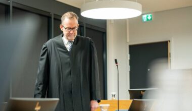 En dommer iført sorte kapper står i en rettssal, med mikrofon og dommerbenk i bakgrunnen. Rommet har moderne interiør med lys og mørke farger. Det er dataskjermer og en glassvase på bordet. Oppsettet skaper en formell atmosfære for rettsbehandlingen.