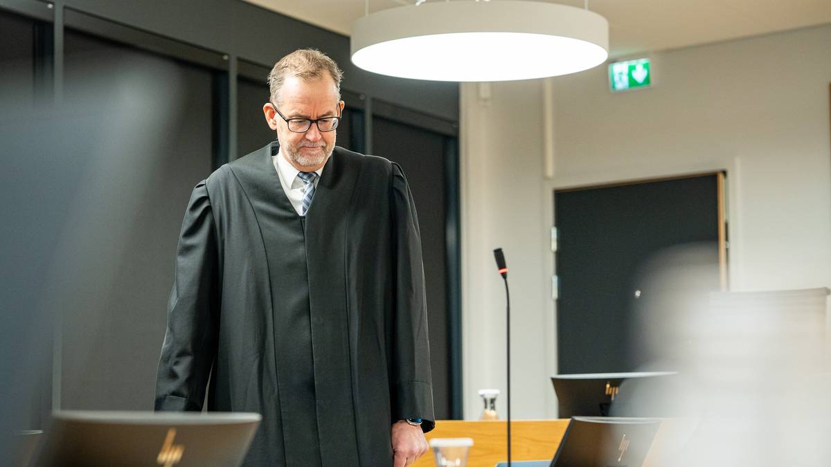 En dommer iført sorte kapper står i en rettssal, med mikrofon og dommerbenk i bakgrunnen. Rommet har moderne interiør med lys og mørke farger. Det er dataskjermer og en glassvase på bordet. Oppsettet skaper en formell atmosfære for rettsbehandlingen.