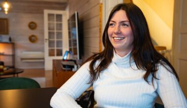 En kvinne sitter ved et bord i et rom med lyse vegger og tregulv. Hun har på seg en lys, stripete genser og har langt, mørkt hår. I bakgrunnen er det en sofa og et TV-apparat, samt en liten spisestue med noen frukter synlig på bordet. Rommet er belyst av naturlig lys som strømmer fra en dør inn til en annen del av boligen. (Bildebeskrivelsen er laget av en KI-tjeneste)