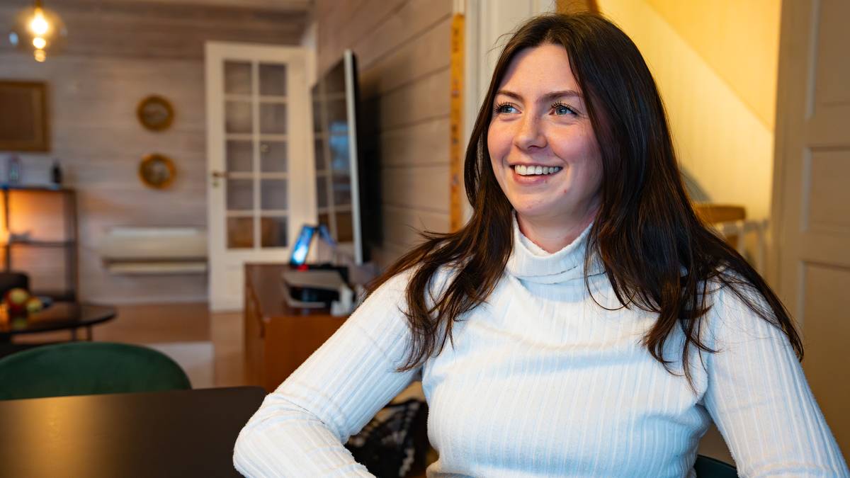 En kvinne sitter ved et bord i et rom med lyse vegger og tregulv. Hun har på seg en lys, stripete genser og har langt, mørkt hår. I bakgrunnen er det en sofa og et TV-apparat, samt en liten spisestue med noen frukter synlig på bordet. Rommet er belyst av naturlig lys som strømmer fra en dør inn til en annen del av boligen. (Bildebeskrivelsen er laget av en KI-tjeneste)