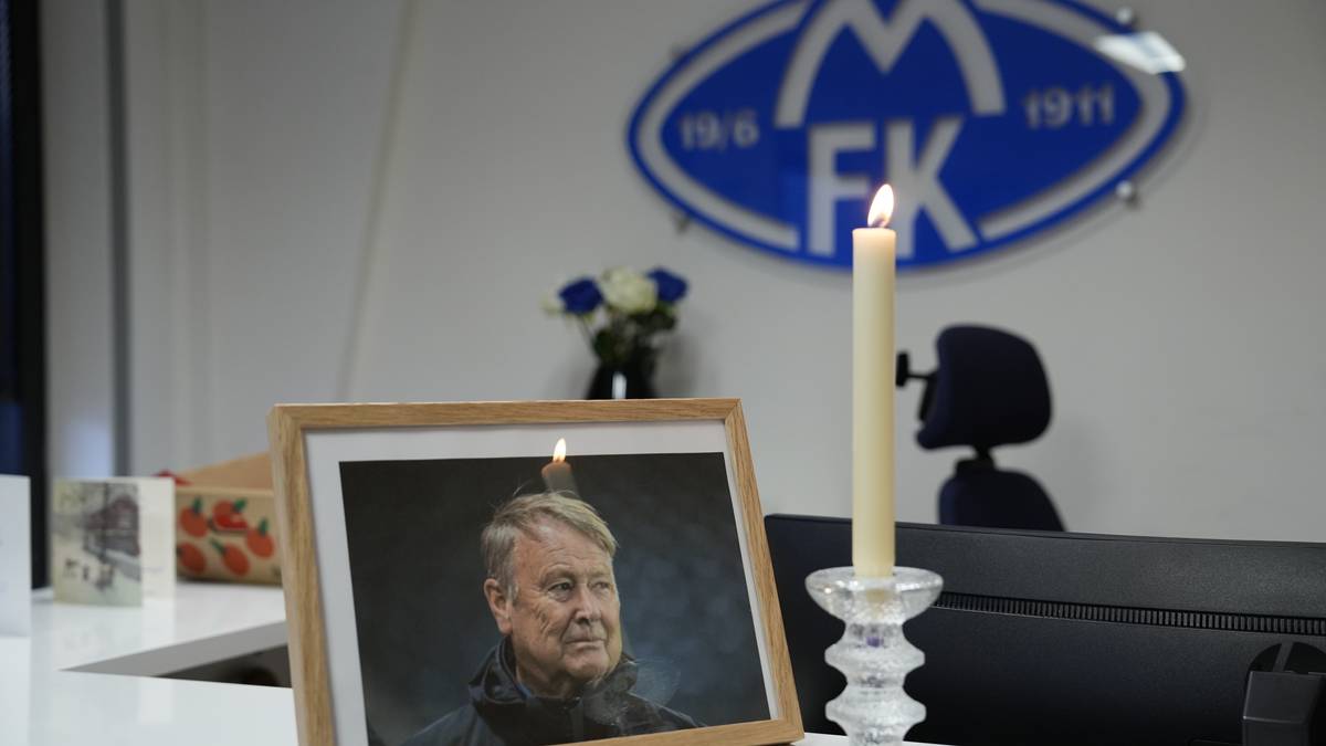 Et bilde viser et bord i et kontorlandskap med et bilde av en person i en ramme, sistnevnte med en tent lys over. I bakgrunnen står en vase med blomster, og i høyre del av bildet sees en kontorstol. Veggen er dekorert med et blått emblem som ser ut til å representere en klubb. (Bildebeskrivelsen er laget av en KI-tjeneste)