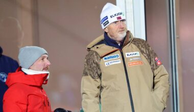 To personer står på en stands i et utendørs arrangement. Den ene er kledd i rød jakke, den andre har på seg en beige jakke. Begge ser mot høyre, muligens mot en pågående aktivitet. Bakgrunnen viser flere personer i vinterklær, noe som indikerer kaldt vær.