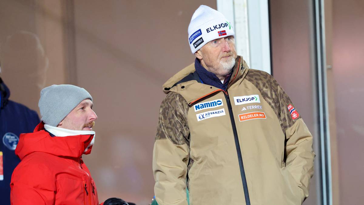 To personer står på en stands i et utendørs arrangement. Den ene er kledd i rød jakke, den andre har på seg en beige jakke. Begge ser mot høyre, muligens mot en pågående aktivitet. Bakgrunnen viser flere personer i vinterklær, noe som indikerer kaldt vær.