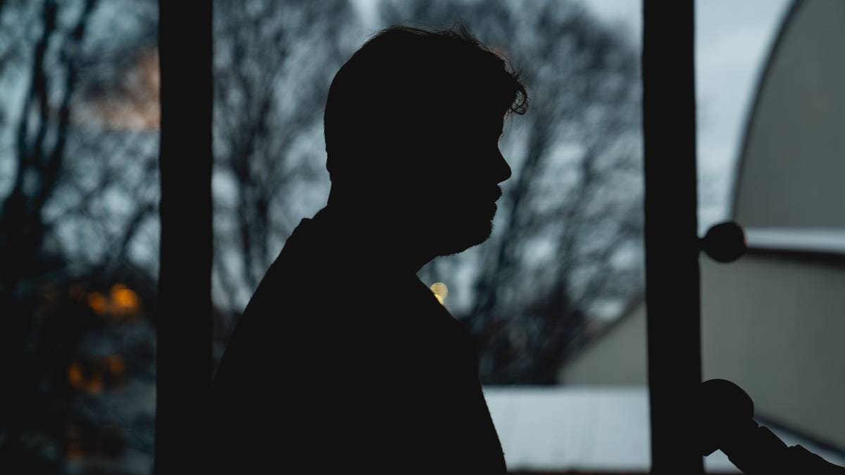 En person står i silhuett foran vinduet, omgitt av en mørk bakgrunn. Det er snø på bakken, og noen lys skinner fra bygninger ute. Scenen gir en stemningsfull atmosfære, trolig i skumringen. Personen virker å være i en refleksiv eller alvorlig tilstand.