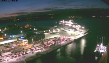 Bildet viser en fergehavn ved skumring, med en stor ferge til kai. Lange kjøretøyslinjer med biler og lastebiler strekker seg mot terminalen. Omgivelsene er belyst av gatebelysning, og det er snø på bakken. I vannet ligger en mindre båt, også opplyst. (Bildebeskrivelsen er laget av en KI-tjeneste)