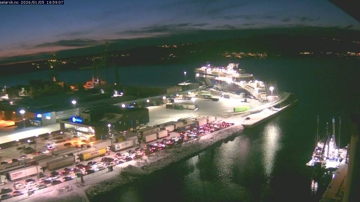 Bildet viser en fergehavn ved skumring, med en stor ferge til kai. Lange kjøretøyslinjer med biler og lastebiler strekker seg mot terminalen. Omgivelsene er belyst av gatebelysning, og det er snø på bakken. I vannet ligger en mindre båt, også opplyst. (Bildebeskrivelsen er laget av en KI-tjeneste)