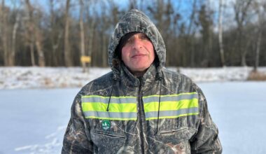 En person står på snødekket underlag iført en camo-jakke med refleksstriper. Bakgrunnen viser trær uten blader, typisk for vinteren. Lyset er klart og himmelen er delvis skyet. Det er ingen synlige objekt i nærheten som distraherer fra personen. (Bildebeskrivelsen er laget av en KI-tjeneste)