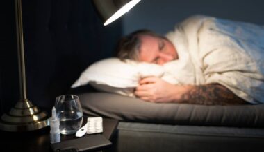 En person ligger til sengs under et teppe, med hender synlig over sengeputen. På nattbordet ved siden av sengen ligger et glass med vann, en pakke med tabletter og en nesespray. Belysningen kommer fra en lampe plassert over sengen. Rommet har dempede farger og en minimalistisk stil. (Bildebeskrivelsen er laget av en KI-tjeneste)