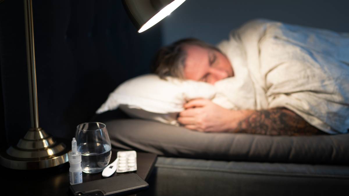 En person ligger til sengs under et teppe, med hender synlig over sengeputen. På nattbordet ved siden av sengen ligger et glass med vann, en pakke med tabletter og en nesespray. Belysningen kommer fra en lampe plassert over sengen. Rommet har dempede farger og en minimalistisk stil. (Bildebeskrivelsen er laget av en KI-tjeneste)