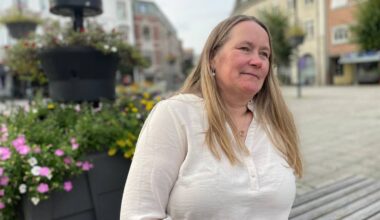 En kvinne sitter på en benk ute. Hun har langt blondt hår, og har på seg en hvit bluse. Hun ser ut mot landskapet.