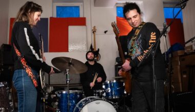 tre musikarar med instrument: gitar, bass, trommer. I eit øvingslokale med lydisolerande matter på veggane.