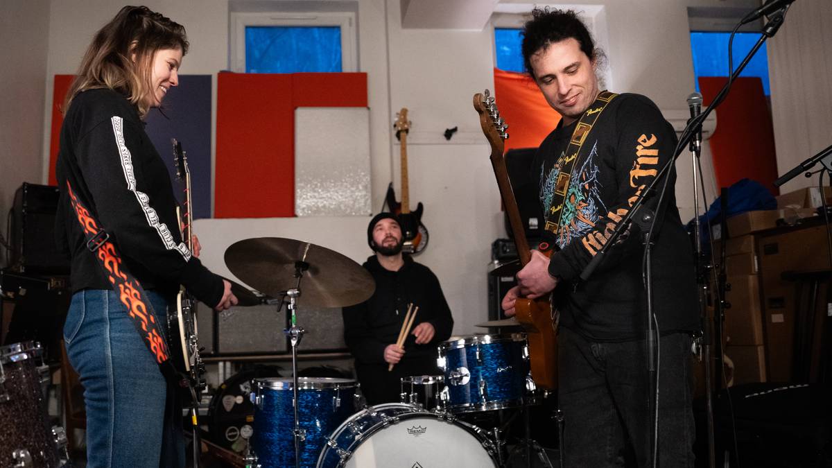 tre musikarar med instrument: gitar, bass, trommer. I eit øvingslokale med lydisolerande matter på veggane.
