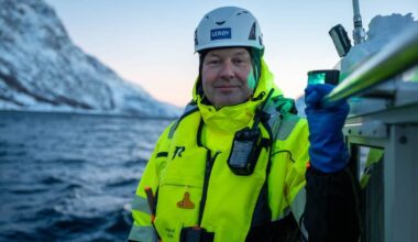 En mann står på en båt med fjell og vann i bakgrunnen. Han har på seg en gul sikkerhetsjakke og hjelm, samt en mobil enhet festet til jakken. Hånden hans holder i et håndtak, mens han holder en grønn lampe i den andre hånden. Belysningen er svak, og det ser ut til å være morgen eller kveld. (Bildebeskrivelsen er laget av en KI-tjeneste)