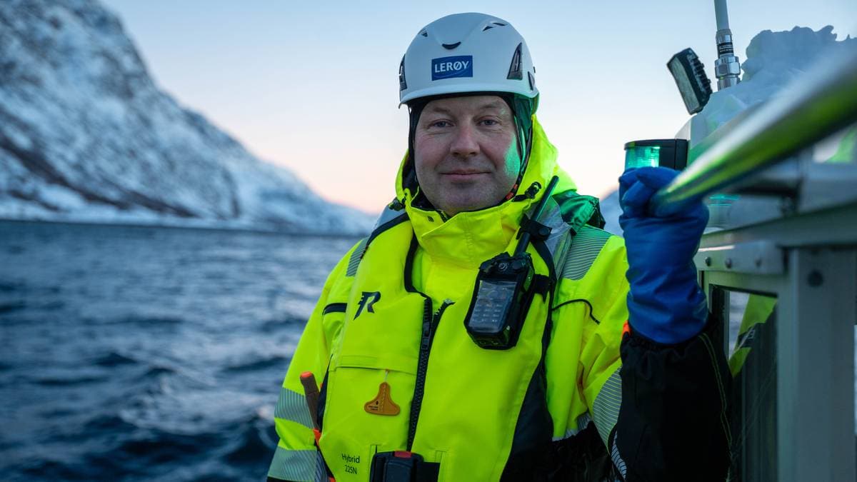 En mann står på en båt med fjell og vann i bakgrunnen. Han har på seg en gul sikkerhetsjakke og hjelm, samt en mobil enhet festet til jakken. Hånden hans holder i et håndtak, mens han holder en grønn lampe i den andre hånden. Belysningen er svak, og det ser ut til å være morgen eller kveld. (Bildebeskrivelsen er laget av en KI-tjeneste)