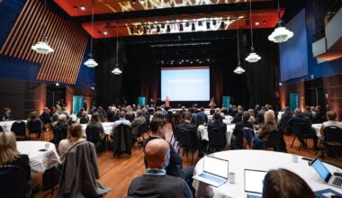 En kvinne står på scenen og holder en presentasjon under en nyttårskonferanse. Publikum sitter rundt bord med hvite duker, og flere deltakere ser mot scenen. Lokalet har mørke vegger med lysinstallasjoner, og det er stoler plassert i en halvsirkel. På scenen er det en stor skjerm med teksten "Nyttårskonferansen 2023". (Bildebeskrivelsen er laget av en KI-tjeneste)