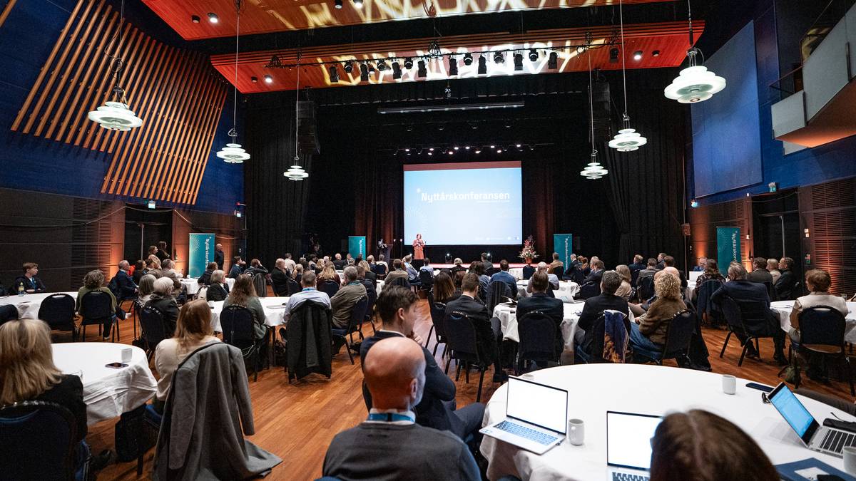 En kvinne står på scenen og holder en presentasjon under en nyttårskonferanse. Publikum sitter rundt bord med hvite duker, og flere deltakere ser mot scenen. Lokalet har mørke vegger med lysinstallasjoner, og det er stoler plassert i en halvsirkel. På scenen er det en stor skjerm med teksten "Nyttårskonferansen 2023". (Bildebeskrivelsen er laget av en KI-tjeneste)