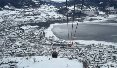 Bildet viser en utsikt fra en taubane over en snødekt by med hytter og bygninger. Det er flere hus med hvite tak og en innsjø som delvis er dekket av is. I forgrunnen ses kabinen til taubanen som er på vei fra bakken. Omgivelsene består av snøfylte landskap og fjellbakgrunn, noe som gir en vinterlig atmosfære.