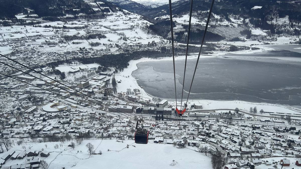 Bildet viser en utsikt fra en taubane over en snødekt by med hytter og bygninger. Det er flere hus med hvite tak og en innsjø som delvis er dekket av is. I forgrunnen ses kabinen til taubanen som er på vei fra bakken. Omgivelsene består av snøfylte landskap og fjellbakgrunn, noe som gir en vinterlig atmosfære.