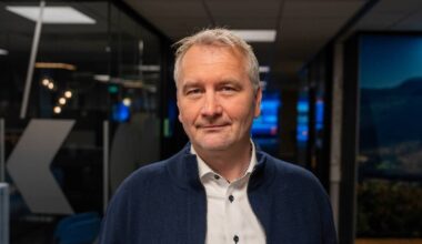 Bildet viser en person kledd i en genser og skjorte, stående i en moderne kontoromgivelse. Bakgrunnen inneholder mørkevegger og skjermer med informasjon synlig i lyse farger. Det er synlige lysekroner og designelementer i rommet som bidrar til et profesjonelt miljø. Belysningen er dempet, noe som gir en alvorlig atmosfære. (Bildebeskrivelsen er laget av en KI-tjeneste)