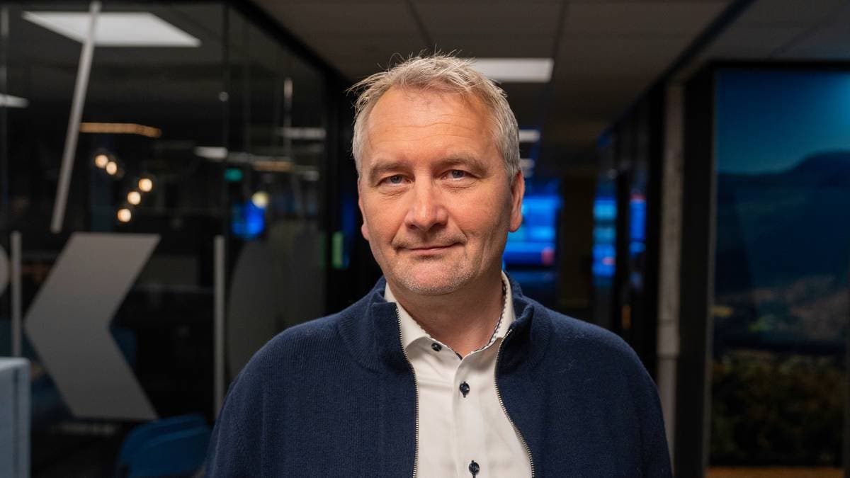 Bildet viser en person kledd i en genser og skjorte, stående i en moderne kontoromgivelse. Bakgrunnen inneholder mørkevegger og skjermer med informasjon synlig i lyse farger. Det er synlige lysekroner og designelementer i rommet som bidrar til et profesjonelt miljø. Belysningen er dempet, noe som gir en alvorlig atmosfære. (Bildebeskrivelsen er laget av en KI-tjeneste)