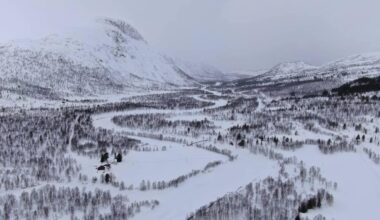 Landskapet viser en snødekt dal med en svingete elv som renner gjennom. Lange, vinterkledde fjell omgir området, og trærne er dekket av snø. I dalen er det et rødt hus synlig blant trærne, omkranset av snødekte marker. Himmelrommet er grått og overskyet, som indikerer kaldt vær.