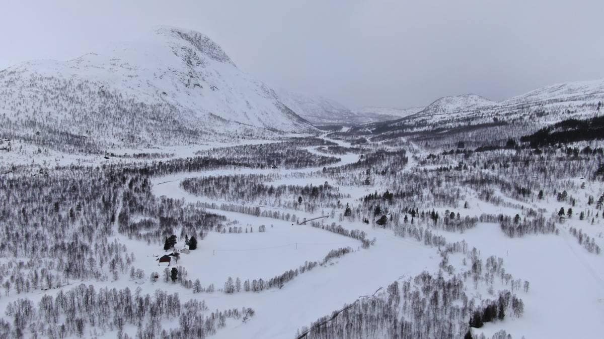 Landskapet viser en snødekt dal med en svingete elv som renner gjennom. Lange, vinterkledde fjell omgir området, og trærne er dekket av snø. I dalen er det et rødt hus synlig blant trærne, omkranset av snødekte marker. Himmelrommet er grått og overskyet, som indikerer kaldt vær.