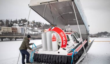 En mann i vinterklær er i ferd med å sikre en redningsbåt på en rampe ved en islagt vannkant. Båten, merket med "SAR," står delvis skjult under et tak. I bakgrunnen er en snødekt by omkranset av trær og bygninger. Det er overskyet, og omgivelsene gir inntrykk av en kald vinterdag. (Bildebeskrivelsen er laget av en KI-tjeneste)