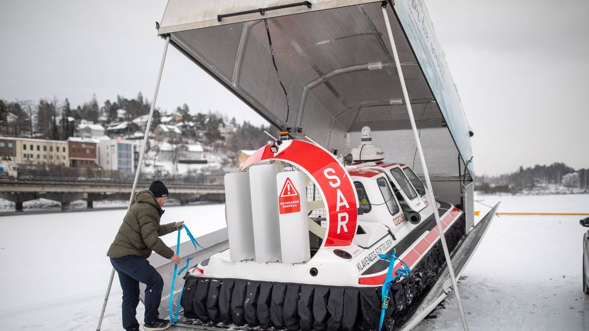 En mann i vinterklær er i ferd med å sikre en redningsbåt på en rampe ved en islagt vannkant. Båten, merket med "SAR," står delvis skjult under et tak. I bakgrunnen er en snødekt by omkranset av trær og bygninger. Det er overskyet, og omgivelsene gir inntrykk av en kald vinterdag. (Bildebeskrivelsen er laget av en KI-tjeneste)